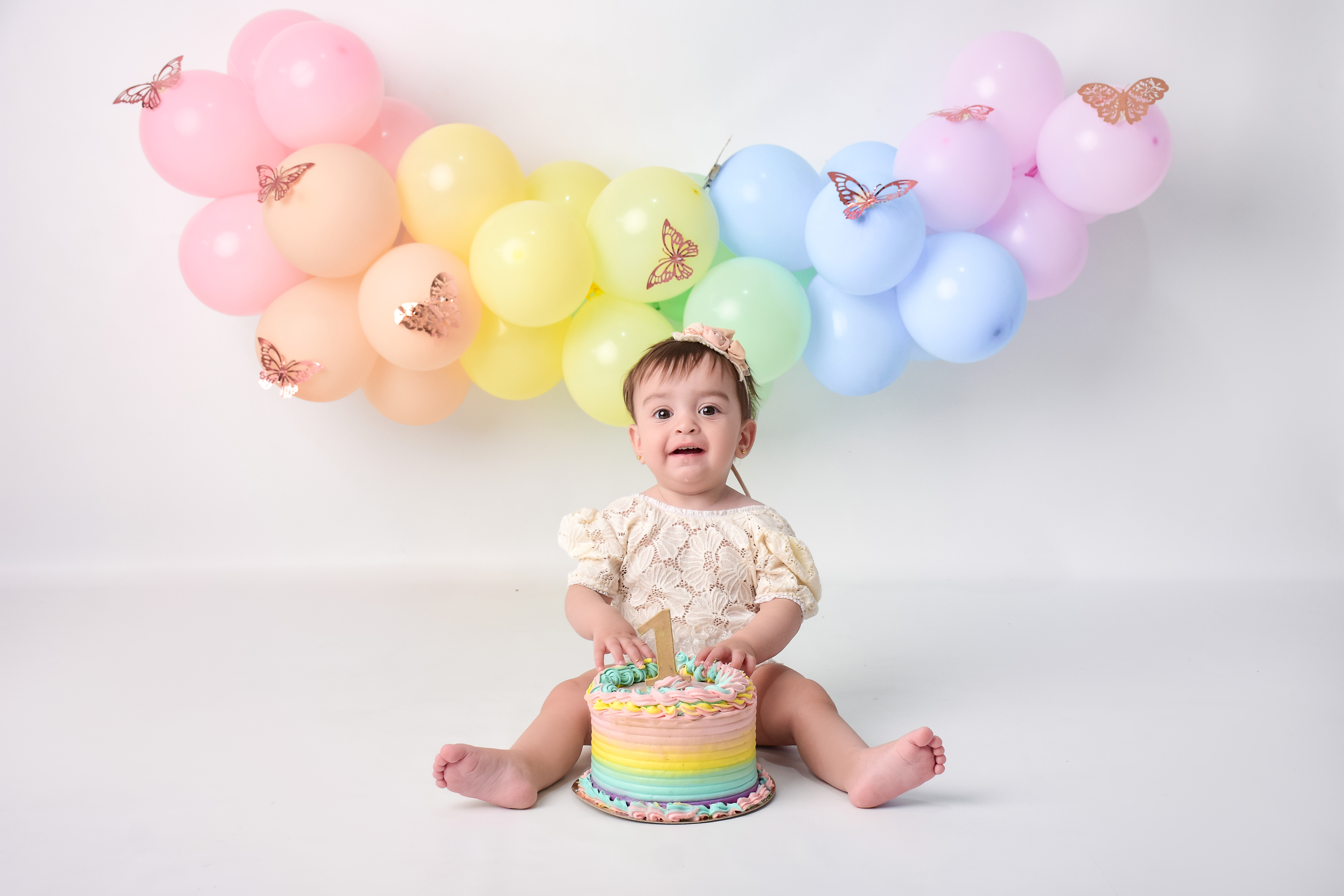 Bebé de un año jugando con una torta arcoíris frente a una guirnalda de globos pastel con mariposas en una sesión de Cake Smash en Lomas del Mirador